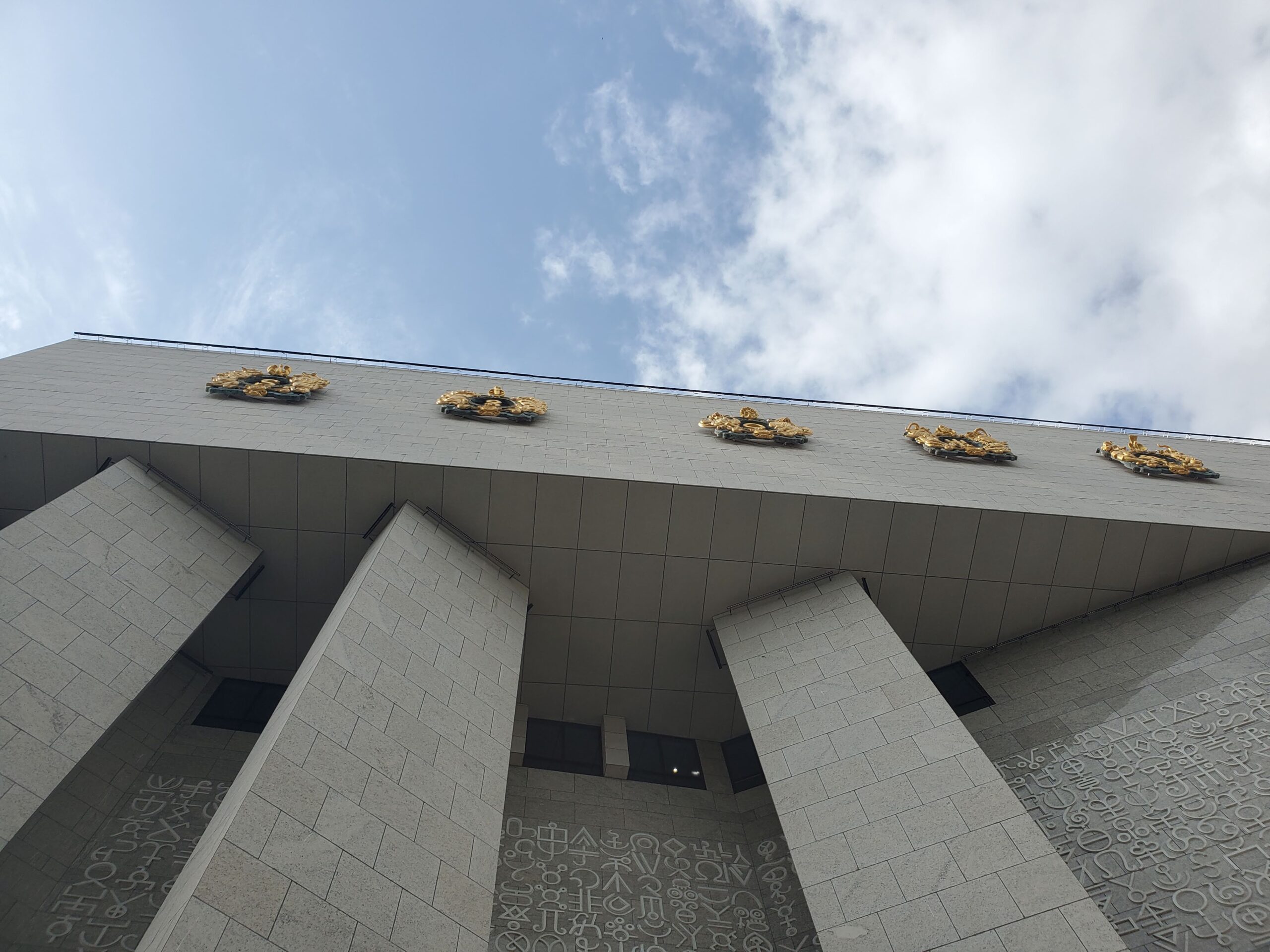 Looking up at a gray stone museum with carvings on the wall and gold images on the facade.