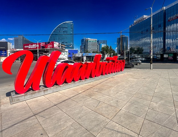 An urban landscape picture featuring a large red red sign reading "Ulaanbaatar" with multiple glass and concrete buildings in the background.