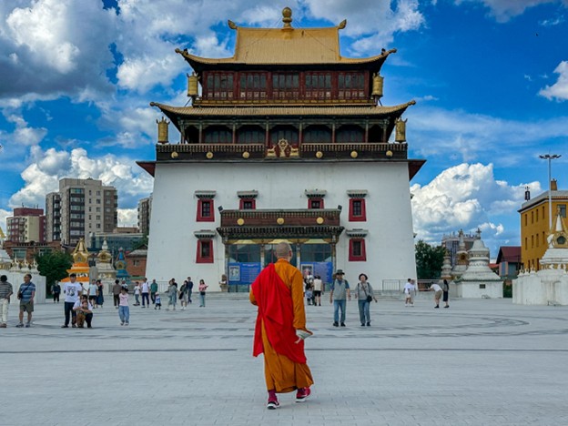 A picture of a Buddhist monk in saffron and red robes walking towards a large white, red, and yellow Buddhist temple.