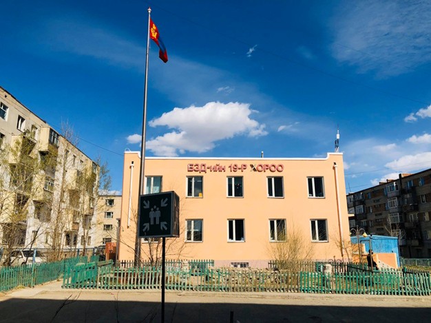 A picture of a small concrete two-story office building with a short green picket fence in front of it and concrete four-story apartment buildings on either side.