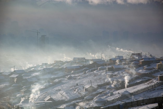 A picture of wooden cabins and white felt gers on a hillside, all of which have thick, white smoke coming from their chimneys that is causing visible smog.