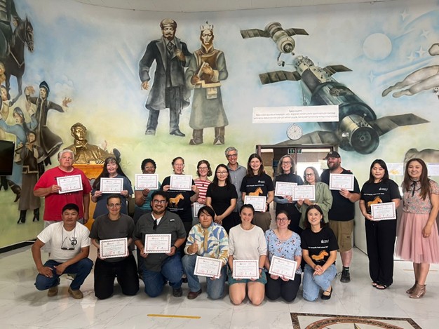 A picture of a group of people ages 18-75, standing in front of a mural holding certificates and smiling at the camera.
