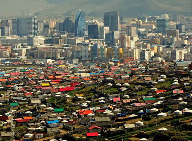 A photo of the sprawling city of Ulaanbaatar, Mongolia, with small gers and hand-built wooden cabins in the foreground and tall steel, glass, and concrete skyscrapers in the background.