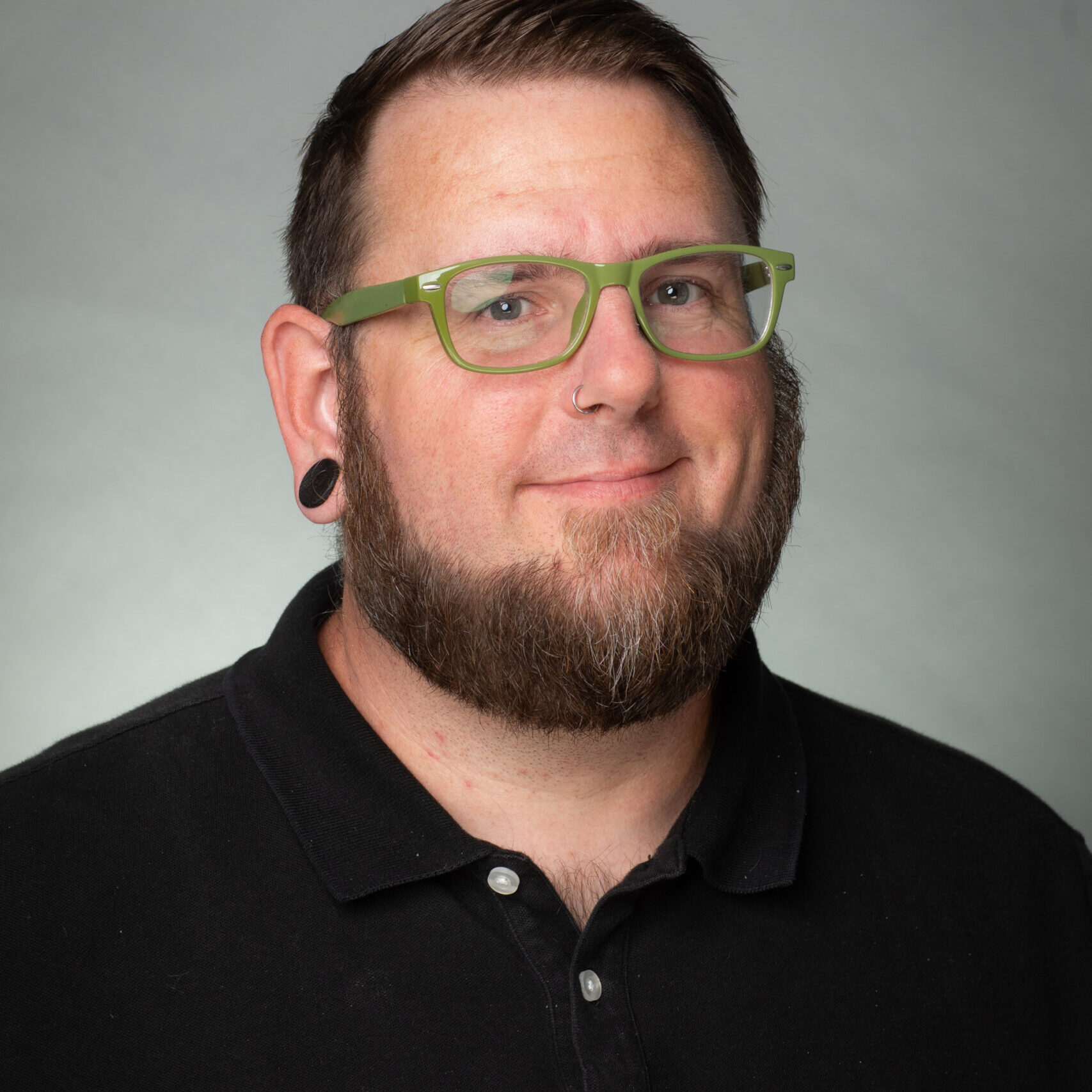 A professional headshot of a white man with brown hair and a beard wearing green glasses and a black polo shirt.