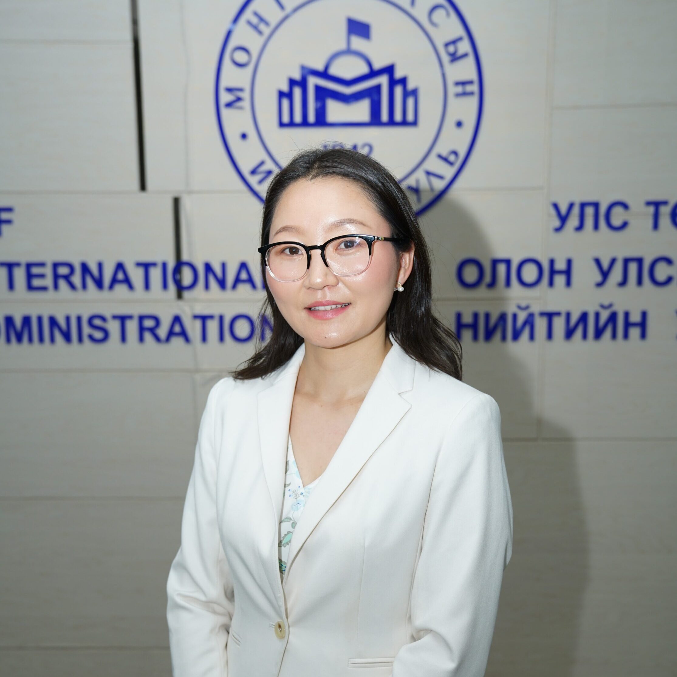 A young Mongolian woman wearing a white suit stands in front of the emblem of the National University of Mongolia and smiles at the camera.