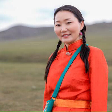 A photograph of a young Mongolian woman standing in a field wearing a bright orange Mongolian garment.
